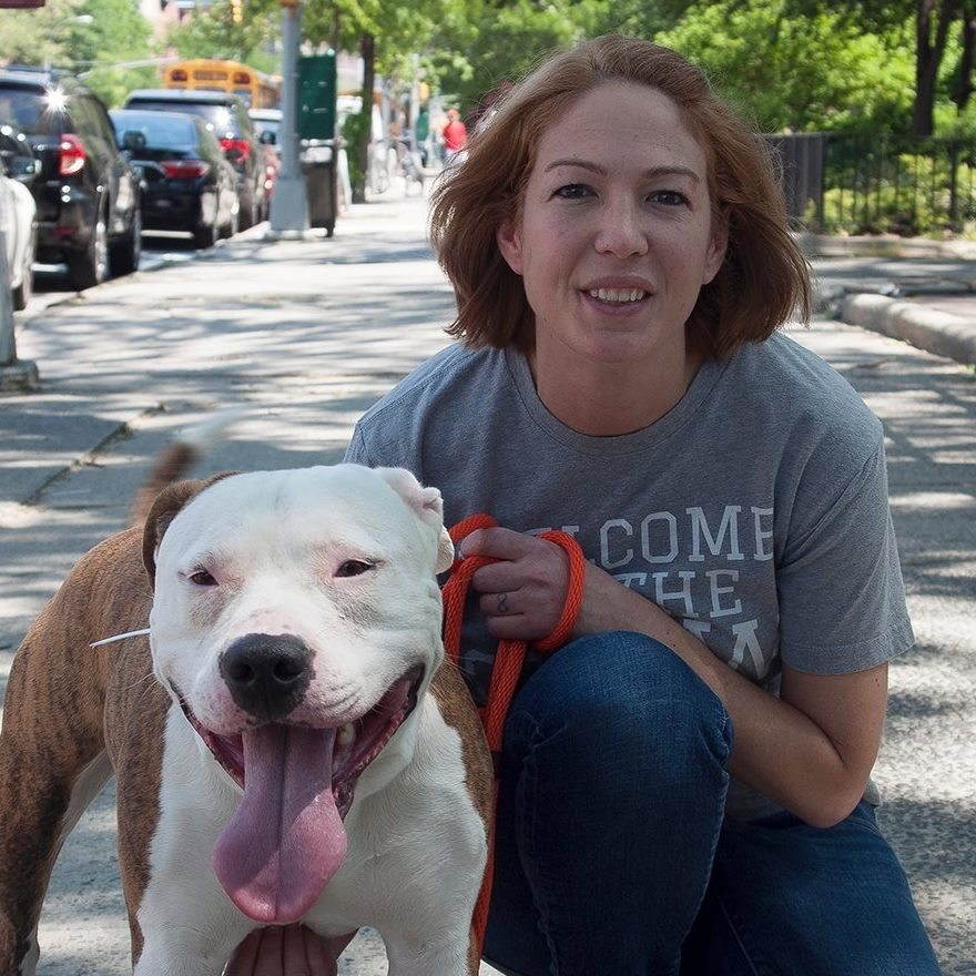 Photo of Kelly Duer, smiling and kneeling next to a white dog on a paved pathway