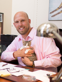 Photo of Brett Frazier at his desk, smiling, wearing a pink dress shirt, holding a gray and white cat