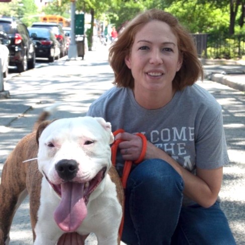 Photo of Kelly Duer, smiling and kneeling next to a white dog on a paved pathway