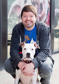 Bio photo of Arlyn Bradshaw, kneeling and smiling in a dark jacket and a green shirt, with a small whie dog in front