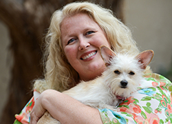 Bio photo of Barbara Camick, smiling in a multicolored shirt, holding a small white dog