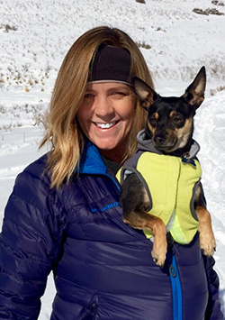 Bio photo of Kathleen Toth, smiling in a snowy winter setting in a blue coat holding a small dog in a yellow coat