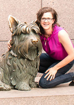 Bio photo of Lisa Gunter, crouching and smiling in a pink shirt and blue jeans by a bronze statue of a scottie dog