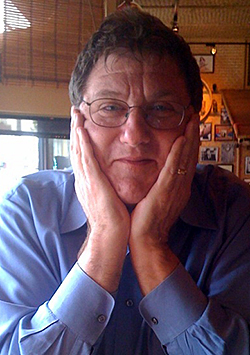 Bio photo of Mark Rovner in a blue dress shirt, smiling, cradling his chin in the palms of his hands