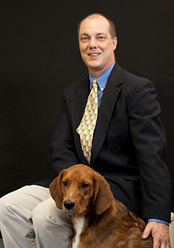 Bio photo of Scott Giacoppo, smiling in a dark suit jacket and khaki pants,sitting next to a reddish dog