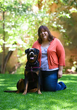 Bio photo of Sue Citro, smiling and kneeling on a lawn with a brown and black dog