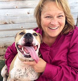 Bio photo of Tawny Hammond smiling in a burgundy shirt in front of a rustic wooden wall with a brown and white dog