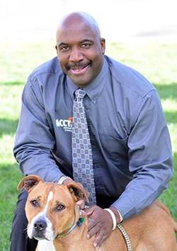 Bio photo of Vincent Medley, kneeling and smiling in a dark gray shirt and tie, petting a brown and white dog