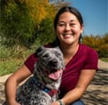 Bio photo of Clare Callison in an outdoor setting, smiling, in a maroon top with a gray dog