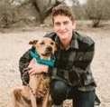 Bio photo of Rory Adams, smiling in an arid outdoor setting kneeling next to a large tan and black dog