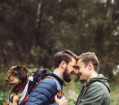 male couple with dog in backpack facing each other in an outdoor setting