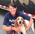 Photo of Colleen Harrington smiling, wearing a blue shirt and kneeling next to a brown and white dog