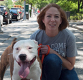 Photo of Kelly Duer, smiling and kneeling next to a white dog on a paved pathway