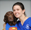 Bio photo of Dr. Kim A. Sanders, smiling in blue scrubs hugging a brown dog in a Superman shirt