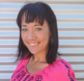 Bio photo of Kristen Auerbach, smiling in a pink shirt against a corrugated metal background