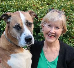 Bio photo of Joyce Briggs, smiling in a black coat over a green top, next to large tan and white dog