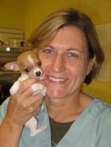 Bio photo of Dr. Natalie Isaza, smiling in light green scrubs, hugging a little brown and white puppy